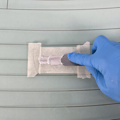 Une main gantée de bleu applique uniformément l'adhésif conducteur argenté MG Chemicals 8331D sur les lignes de masquage d'une lunette arrière, dans le cadre de la réparation du dégivreur. Cet adhésif rétablit la conductivité électrique de la zone endommagée.