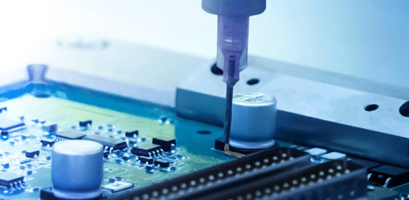 Automated dispensing needle applying adhesive to the edge of an integrated circuit on a printed circuit board during an electronics manufacturing process.
