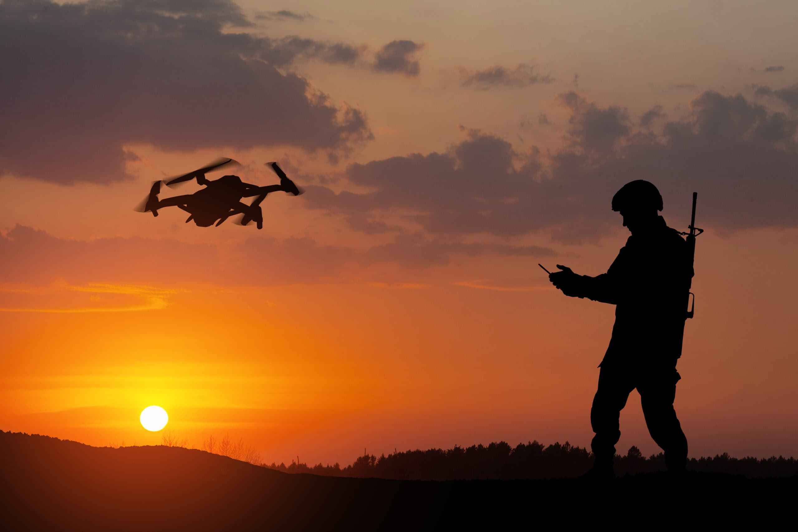A silhouetted soldier operates a handheld controller while a quadcopter drone flies nearby at sunset, with an orange sky and low clouds above a dark landscape.