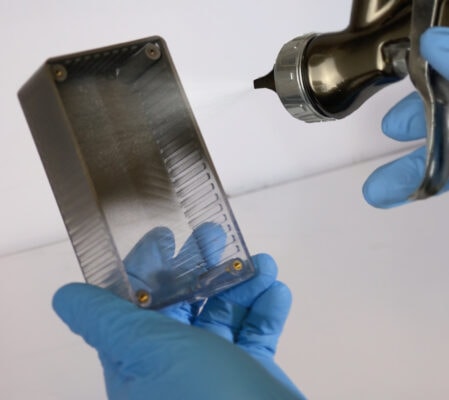 A gloved hand holds a small plastic enclosure while a spray nozzle applies a UAV coating to the interior surface.