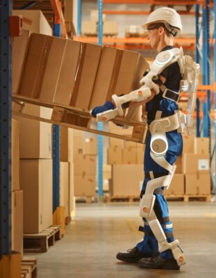 Warehouse worker wearing a powered exoskeleton lifts a pallet of cardboard boxes from a storage rack inside a large warehouse.