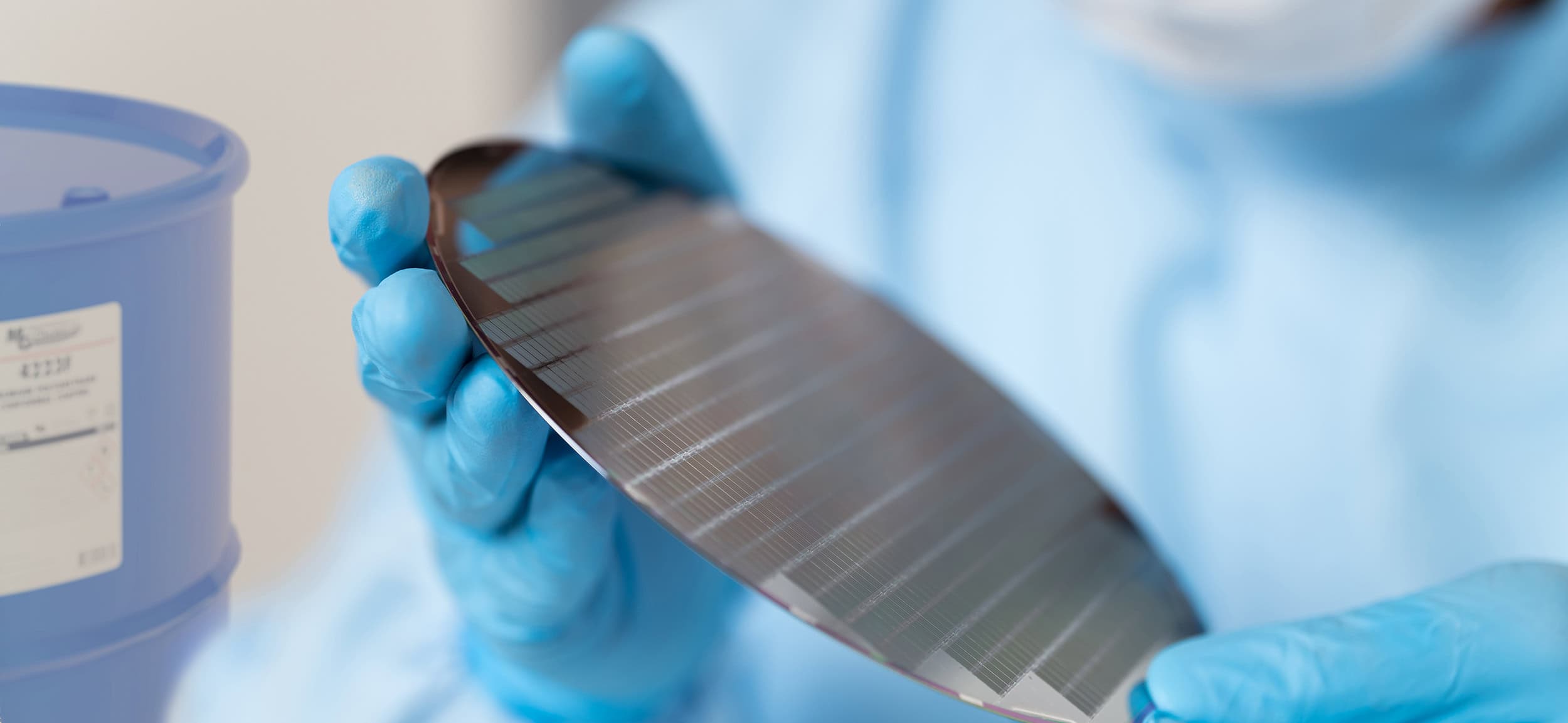 Gloved hands holding a thin, flexible electronic wafer or film in a laboratory setting, with a chemical container visible in the background.