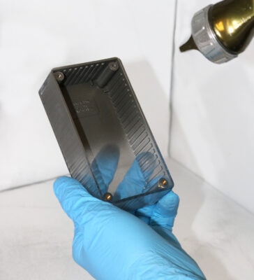 Person wearing blue nitrile gloves spray-coating a rectangular electronic enclosure with EMI shielding paint using a spray gun, applying conductive coating to enhance electromagnetic shielding inside the housing.