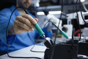 Un técnico sostiene un soldador en un banco de trabajo electrónico, con herramientas y cables de soldadura en primer plano y equipo de laboratorio desenfocado al fondo. La imagen tiene poca profundidad de campo, enfocando la mano y el mango de la herramienta durante un trabajo de reparación o ensamblaje electrónico.