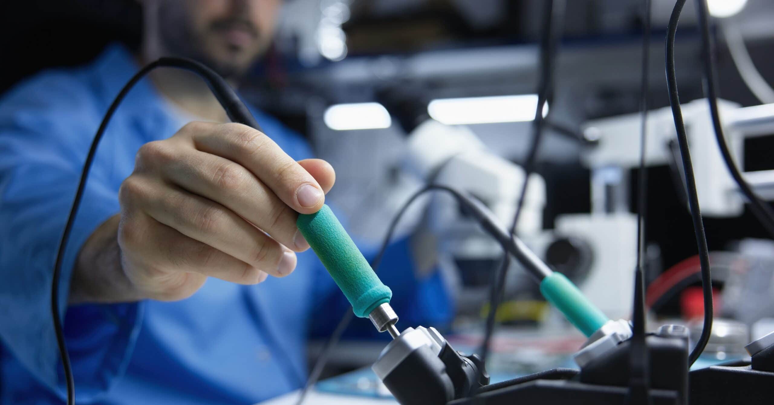 A technician holding a soldering iron at an electronics workbench, with soldering tools and wires in the foreground and lab equipment blurred in the background. The image has shallow depth of field, focusing on the hand and tool handle during electronic repair or assembly work.